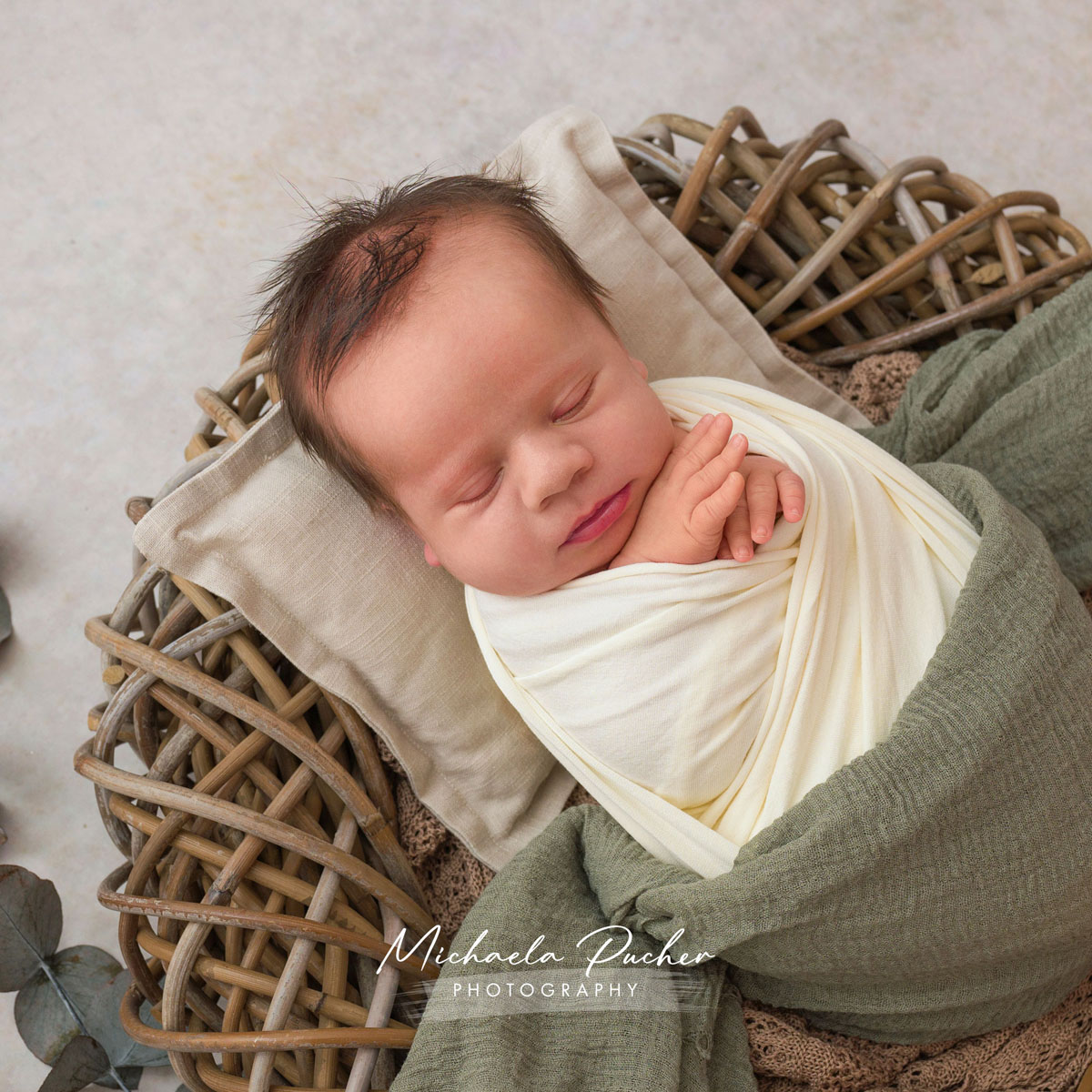 Schlafendes Neugeborenes liegt eingewickelt in cremefarbenen und grünen Tüchern in einem geflochtenen Korb und ruht auf einem kleinen Kissen.